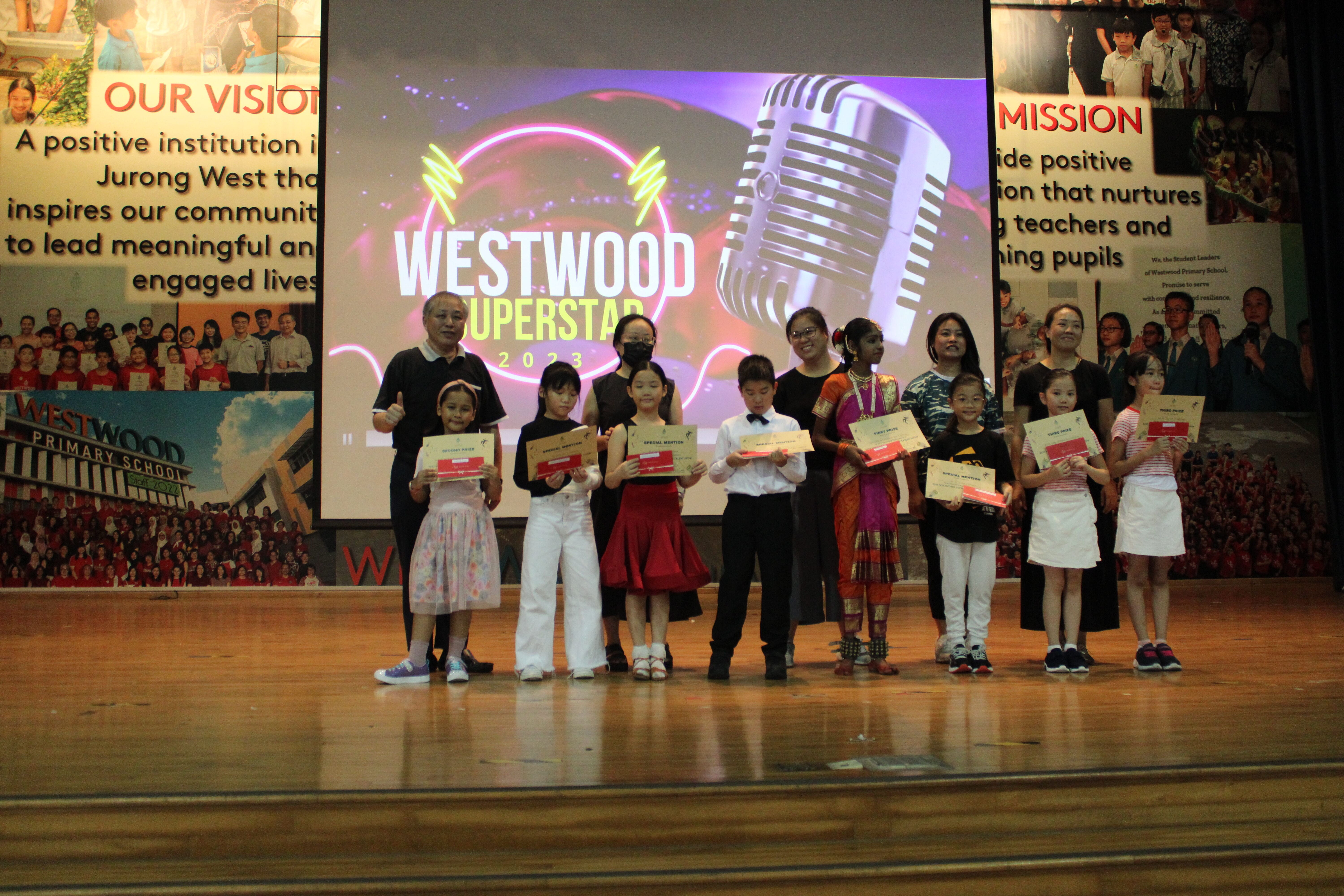 Children and adults stand on a school stage holding certificates at the Westwood Superstar 2023 event.